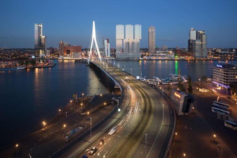 A cityscape at dusk featuring the modern cable-stayed bridge near nhow Rotterdam, surrounded by tall buildings and illuminated streets along the river.