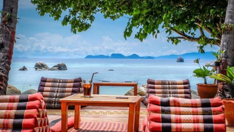 Low wooden tables and cushioned seating at Ten Moons Koh Lipe are arranged under trees on a beach, facing calm blue water and distant islands, with a boat visible on the horizon.