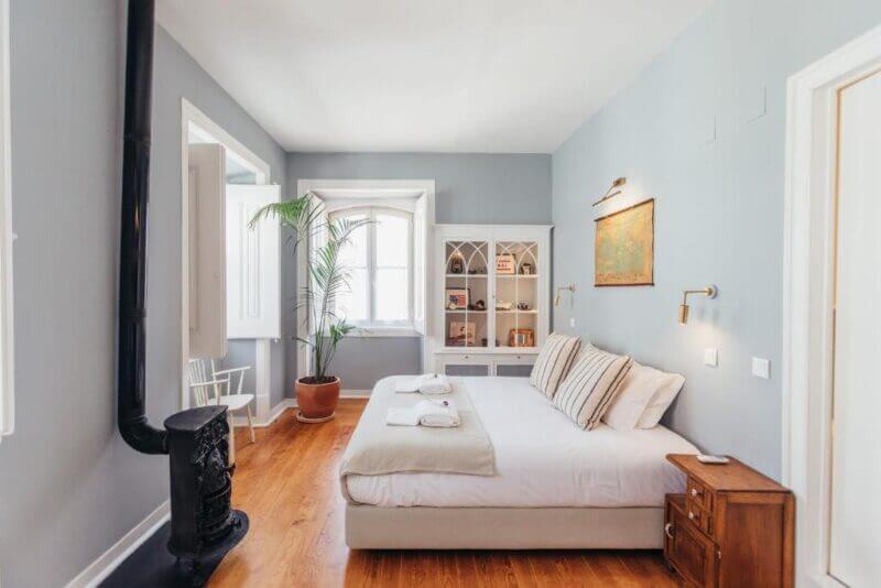 A tidy bedroom in Cascais with a double bed, wooden floor, black wood stove, bookshelf, potted plant by the window, and light blue walls exudes cozy Legasea charm.