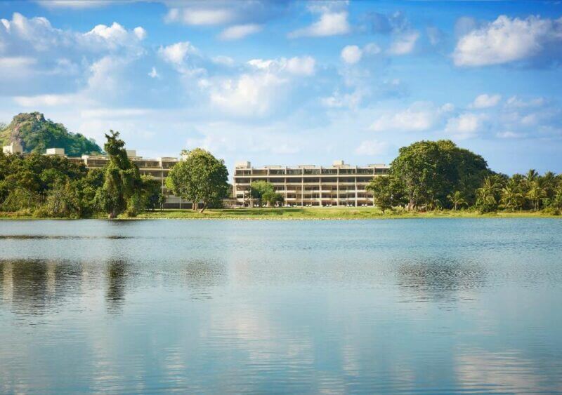 The Jetwing Dambulla building complex stands behind trees on the far side of a calm lake, with a partly cloudy sky and a small hill in the background.