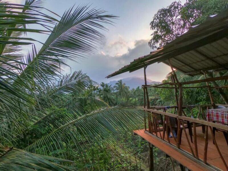 Open-air wooden patio with a checkered table overlooks lush green tropical foliage and distant Blue Mountains under a cloudy sky at sunset.
