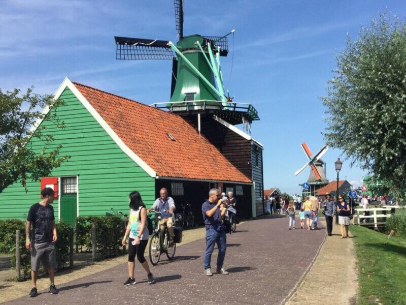 People walk and cycle along a path beside green wooden buildings and traditional windmills on a sunny day, enjoying the scenic views near Zaan-Heerlijck-Slaapen.