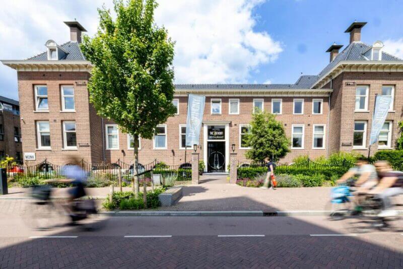 Brick building with banners at entrance, trees and shrubs in front, and several people riding bicycles along the street in daylight near anthony-utrecht.