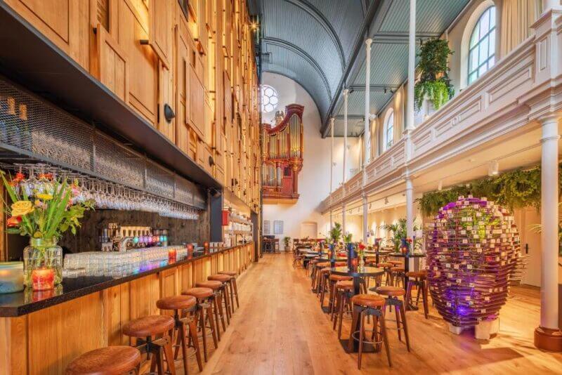 A spacious bar and restaurant inside a converted church, Bunk-Utrecht features high arched ceilings, a grand pipe organ, wooden tables and stools, and vibrant decorative elements.