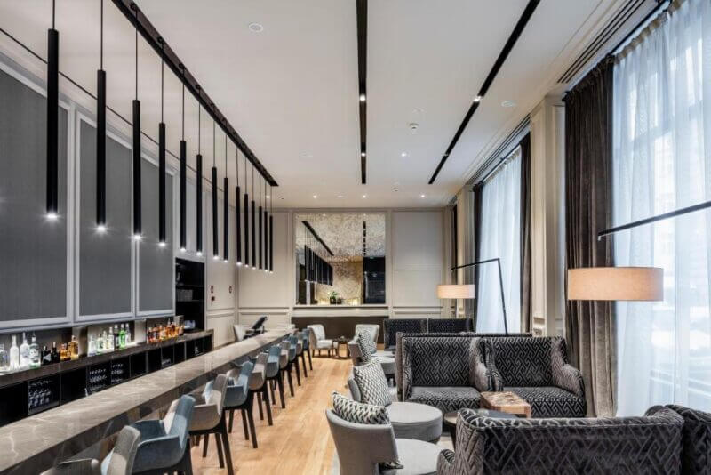Chic Boho hotel lounge in Prague featuring a long bar with high stools, cozy armchairs and sofas, expansive windows with curtains, and stylish pendant lighting overhead.