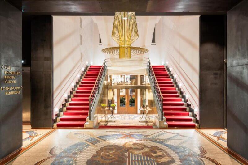 A grand entrance hall in a mosaic house in Prague features two red-carpeted staircases, a large gold chandelier, and glass doors leading to another room.