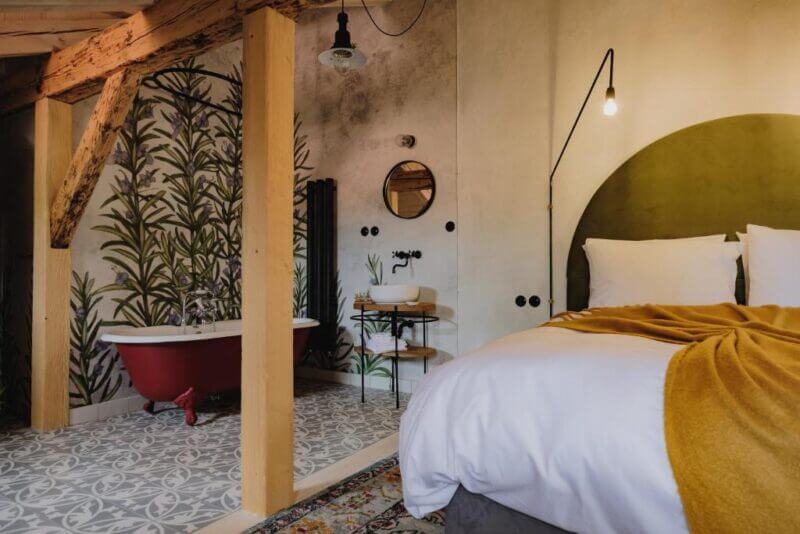A cozy bedroom with a large bed, yellow blanket, clawfoot bathtub, patterned floor tiles, and leafy wall design under exposed wooden beams—evoking the tranquility of a Cesky Krumlov monastery garden.