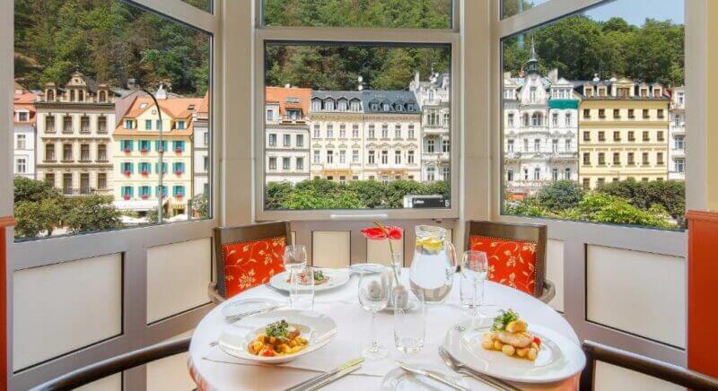 A round dining table set for two with plated meals, glassware, and flowers by a large window overlooks the colorful historic buildings and lush greenery of Karlovy Vary outside.