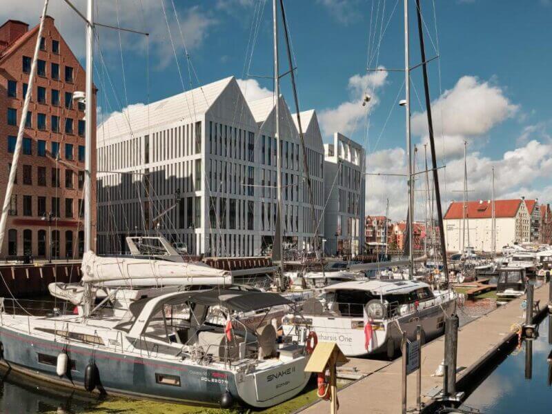 Modern sailboats docked at a marina with contemporary white buildings, historic brick structures, and renowned accommodation Gdansk options in the background under a partly cloudy sky.