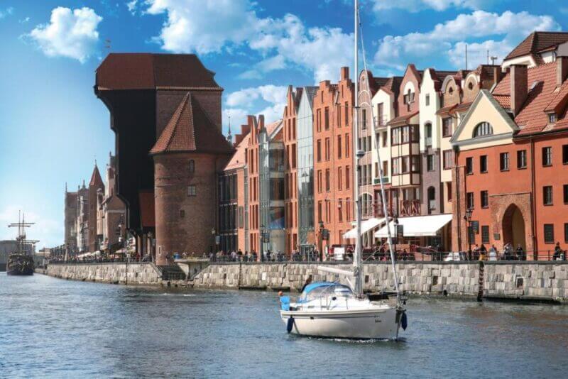 A sailboat glides along a canal in hanza-gdansk, passing historic brick buildings and a medieval tower beneath a blue sky dotted with clouds.