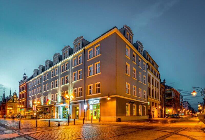 A corner view of the modern, four-story Korona Hotel in Wroclaw, with illuminated windows and outdoor lights casting a glow over the cobblestone street at dusk.