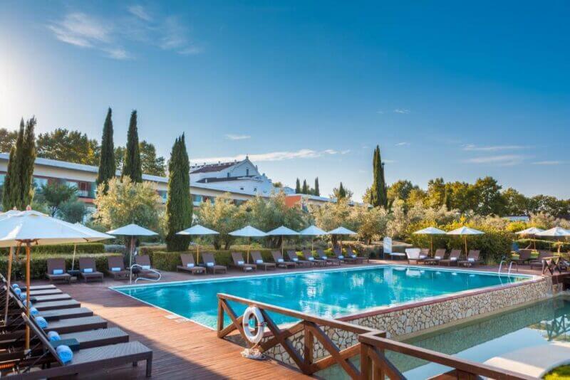Outdoor swimming pool surrounded by lounge chairs and umbrellas, set amid trees and a modern building at the elegant Convento Hotel under a clear blue sky.