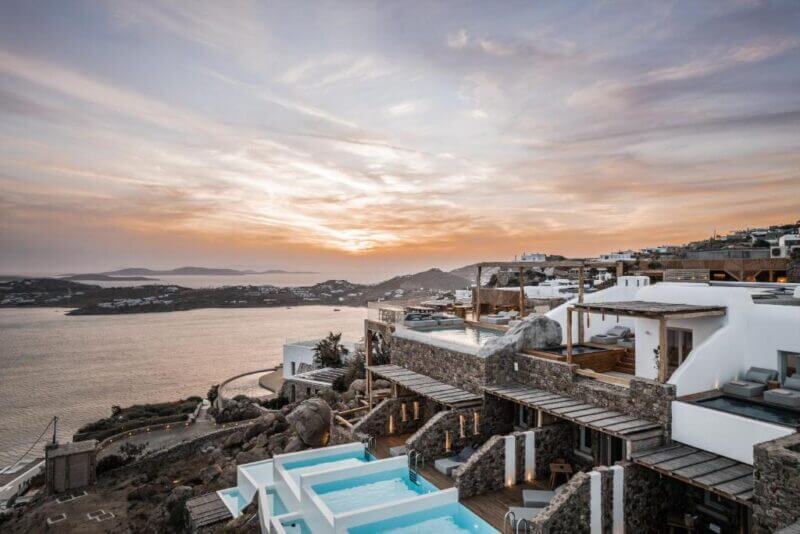 Alissachni Mykonos is a coastal resort with white buildings and private pools overlooking the sea at sunset, with islands and a winding road visible in the distance.