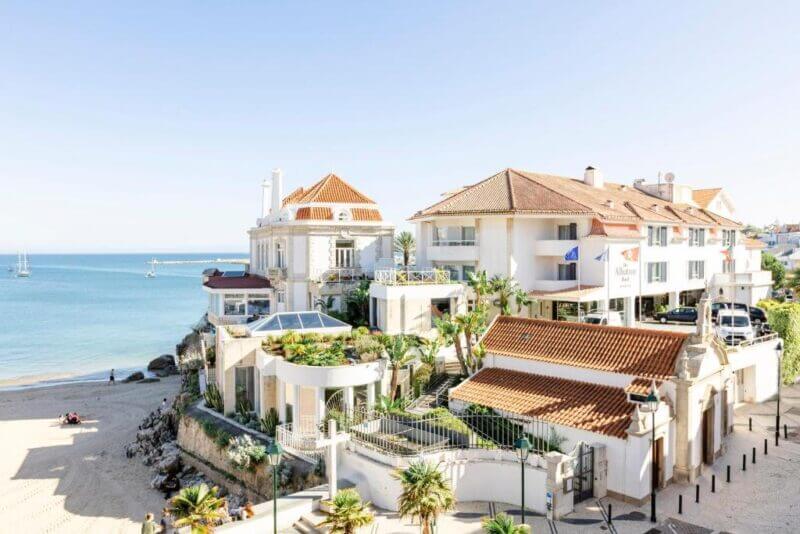 Seaside buildings with red-tiled roofs overlook a sandy beach and calm blue ocean under a clear sky in Cascais; a few people are on the beach, boats are visible in the distance, and the iconic Albatroz hotel stands nearby.