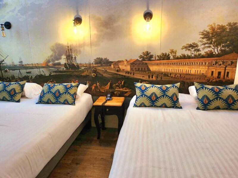 Two beds with patterned pillows in a room with white bedding at Rochefort Hotel, featuring a wall mural depicting a historical scene with ships and buildings.