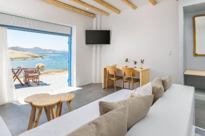 Bright room with white walls, wooden furniture, and a sea view through open double doors. This inviting Milos beach suite features a small table with chairs near the TV and outdoor seating on a patio by the water.