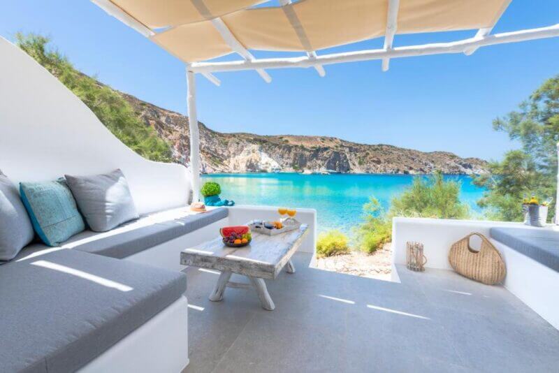 Outdoor patio at Milinon Suites hotel on Milos, with white furniture and a wooden table set with drinks and fruit, overlooking the clear blue sea and rocky coastline under a sunny sky.