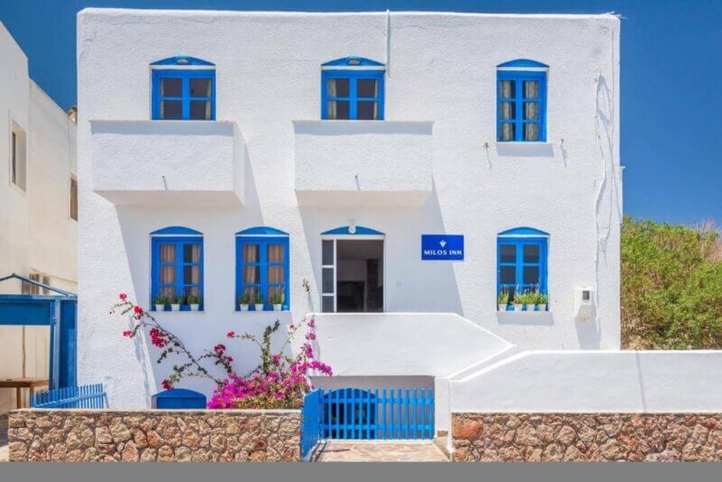 A white two-story building with blue window frames and doors, labeled Milos Inn, featuring a small garden and stone wall in front under a clear blue sky.