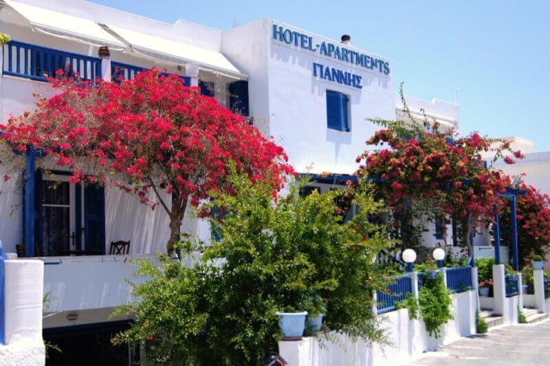 White building with blue accents labeled Giannis-milos Hotel-Apartments, adorned with vibrant pink bougainvillea flowers and greenery under a clear blue sky.