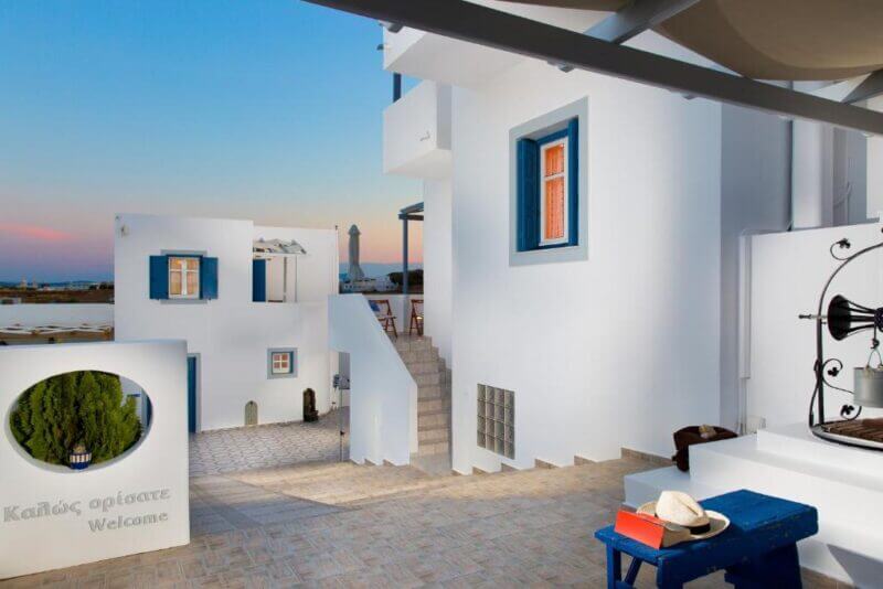 Courtyard of a white Vivere-milos building with blue window frames, outdoor staircase, potted plants, and a bench with a hat and bag, all bathed in the warm glow of sunset.