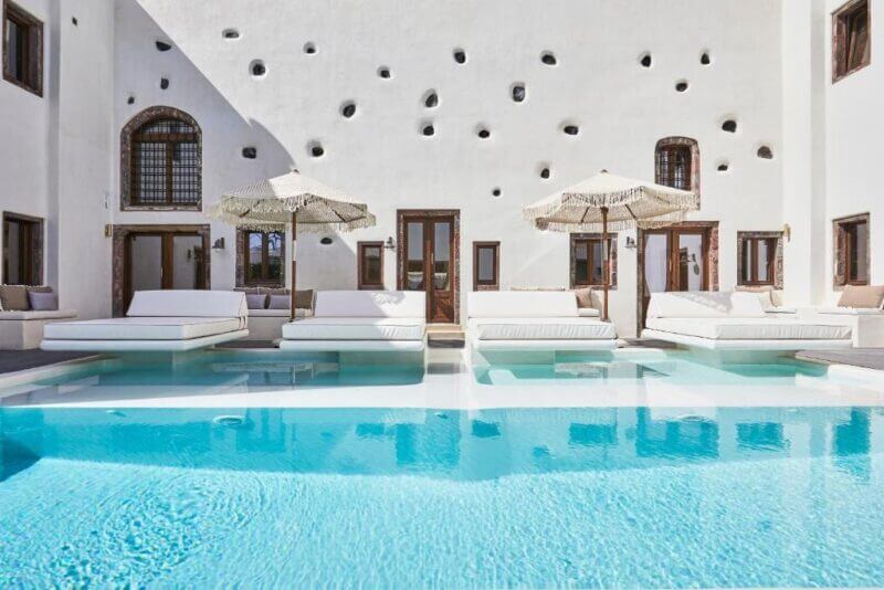 Outdoor pool with clear blue water, surrounded by white lounge beds and umbrellas, set against a white Santorini hotel building with small circular wall openings and wooden doors—evoking the charm of a luxurious winery retreat.