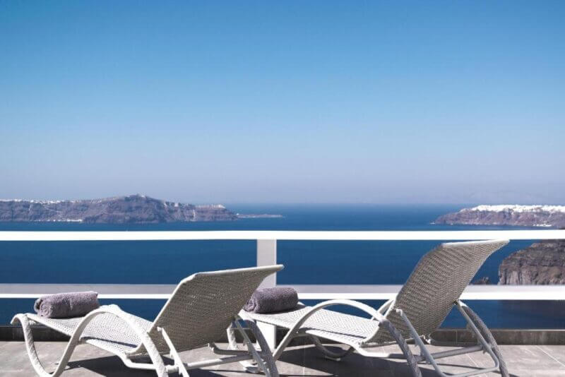Two lounge chairs with towels face a blue sea and distant islands, positioned on a balcony with a white railing under a clear sky at the elegant Santorini hotel Thireas.