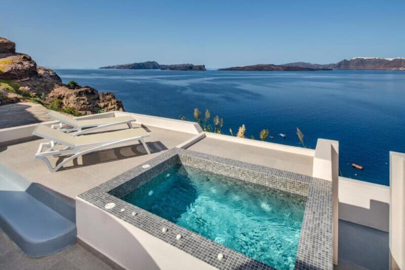 Outdoor hot tub and white benches on a patio at a Santorini hotel, overlooking a calm blue sea and distant rocky islands under a clear sky.