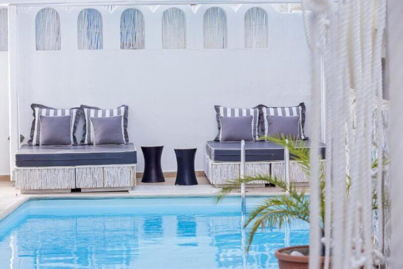 Two cushioned lounge chairs with striped pillows and two small black tables are positioned beside a clear blue swimming pool at Loizos Santorini Hotel, with a white wall and potted plant in the background.