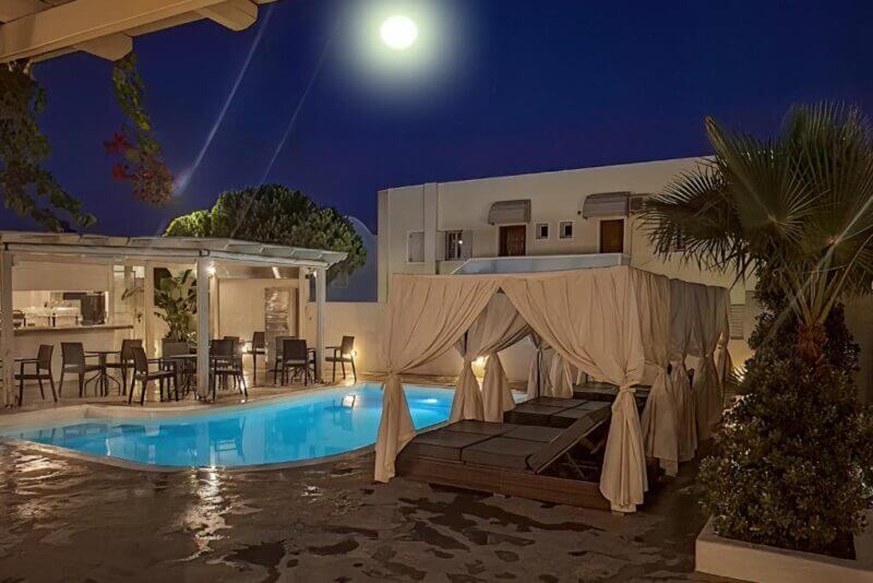 A night view of the poolside lounge at Ambience Suites, Santorini, with curtained cabanas, chairs, tables, and palm trees softly illuminated by moonlight.