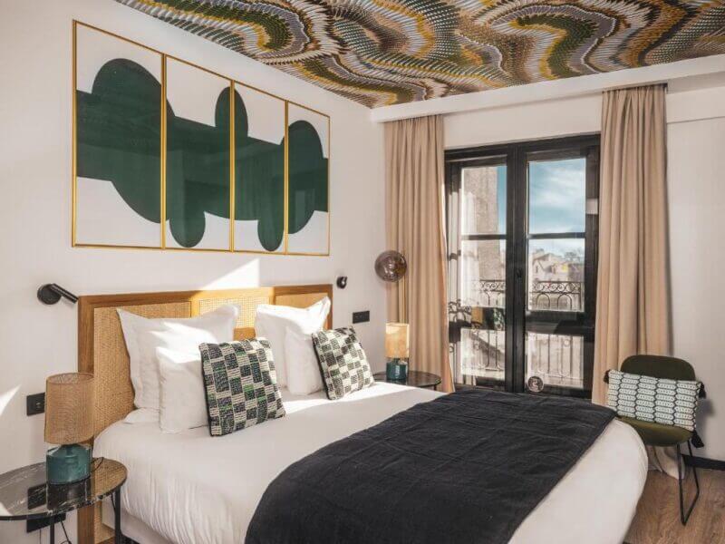 Modern hotel room in La Rochelle featuring a double bed, patterned pillows, wall art, glass side tables, and floor-to-ceiling windows with beige curtains and a decorative patterned ceiling—perfect for your Monde getaway.