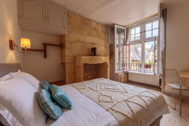 A cozy bedroom at le-lys-sarlat with a double bed, two blue cushions, a decorative fireplace, wooden shelves, a desk and chair, and an open window offering rooftop views.