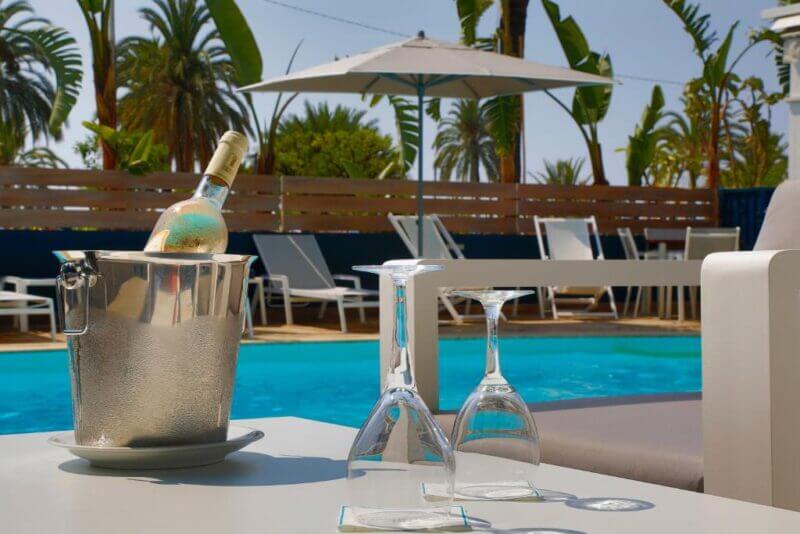 Two empty wine glasses and a chilled bottle of white wine in an ice bucket sit on a table beside the pool at the Napoleon Hotel in Menton, with lounge chairs, palm trees, and an umbrella in the background.