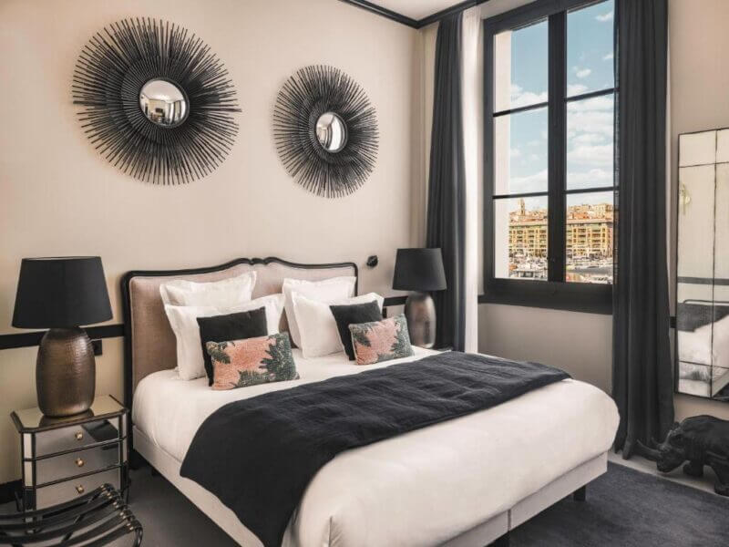 Modern bedroom with large bed, black-and-white decor, two circular wall art pieces, bedside lamps, and a window showing buildings and blue sky—ideal Marseille accommodation for a stylish stay.
