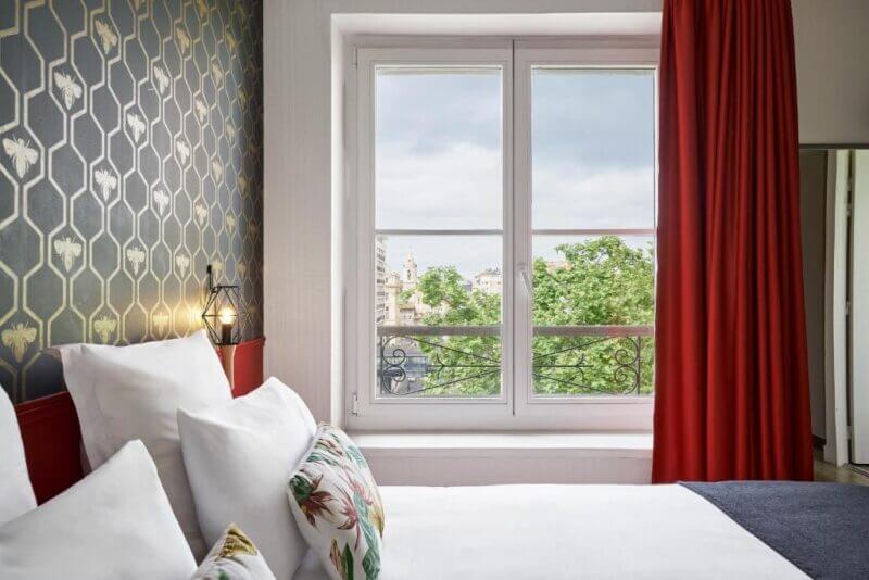 A neatly made bed at a BW Hotel in Marseille sits next to a large window with red curtains, patterned wallpaper, and a view of trees and buildings outside.