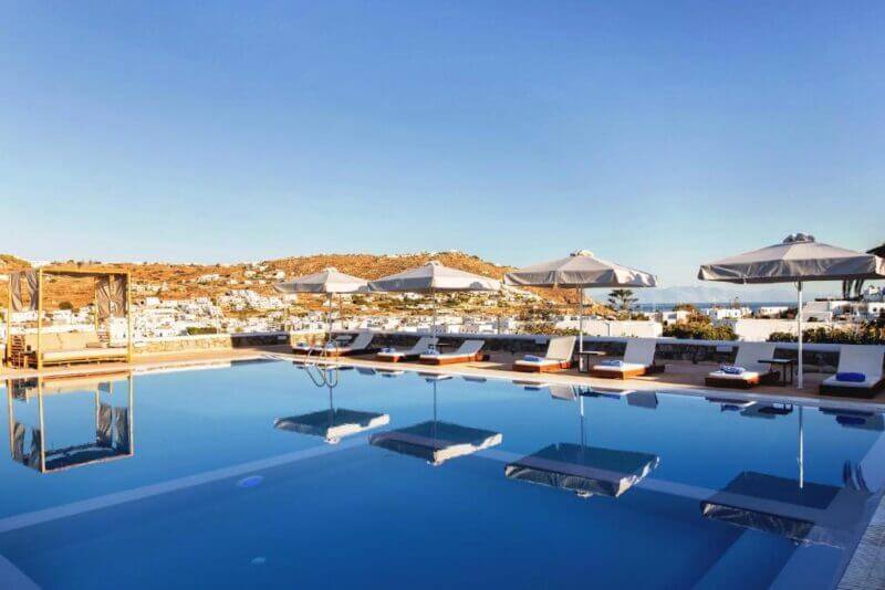 Outdoor swimming pool with sun loungers and umbrellas, overlooking a hillside town and clear blue sky—a stunning feature of Osom Mykonos, your choice for luxury accommodation in Mykonos.