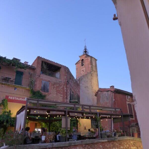 Outdoor restaurant with string lights in front of rustic, weathered maisons and a clock tower at dusk under a clear Roussillon sky.