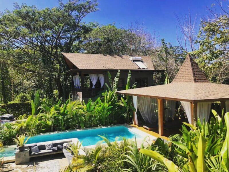 A modern house with a pool and gazebo surrounded by lush tropical plants and trees under a clear blue sky, capturing the relaxed elegance of Lua Villas in Santa Teresa.
