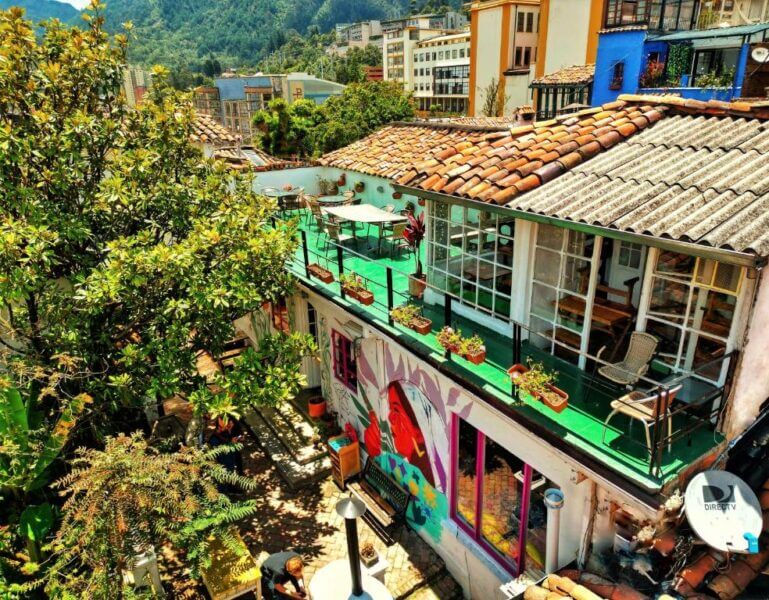 Colorful outdoor café with a mural, potted plants, and tables on a balcony at Botanico Hostel, surrounded by trees and buildings in the hilly urban area of Bogota.
