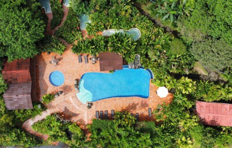 Aerial view of LaFortuna CasaLuna's rectangular outdoor swimming pool and smaller round pool, surrounded by lush greenery, brick pathways, lounge chairs, and shaded seating areas.