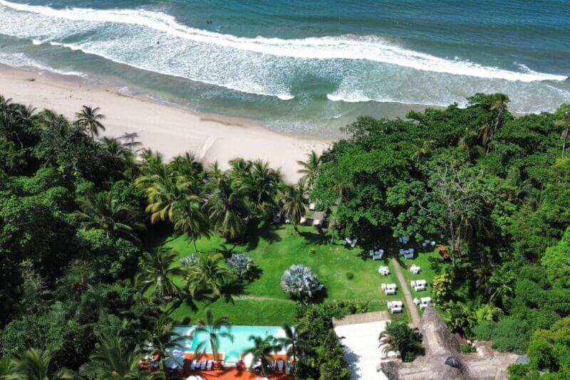 Aerial view of Le Cameleon, a beachfront property in Puerto Viejo with a lush lawn, swimming pool, scattered tables, dense tropical trees, and waves gently rolling onto the sandy shore.