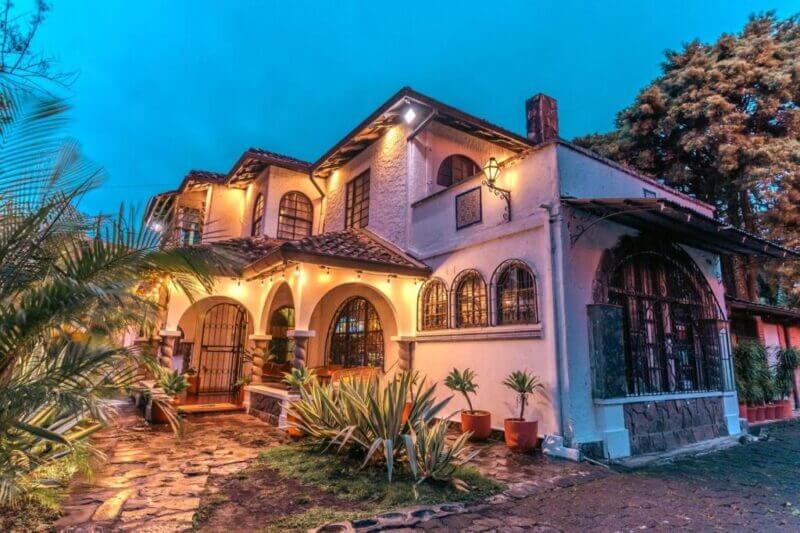 Two-story house with arched windows and warm exterior lighting, surrounded by plants and stone walkway, photographed at dusk in Finca Escalante, San Jose—an inviting hostel atmosphere.