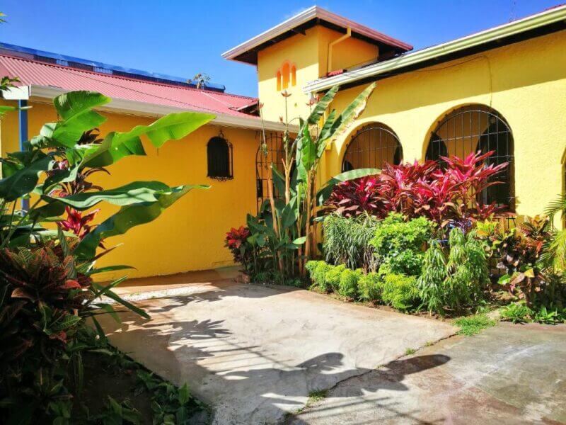 A yellow house in San Jose with arched windows, a red roof, and a small garden filled with tropical plants in the courtyard under a clear blue sky—welcome to Casa 69.