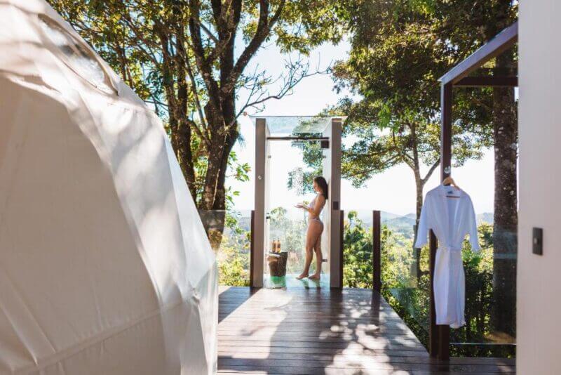 A person stands on a wooden deck surrounded by trees at Chira Glamping in Monteverde, next to an outdoor shower and a robe hanging nearby, with a tent structure to the left.