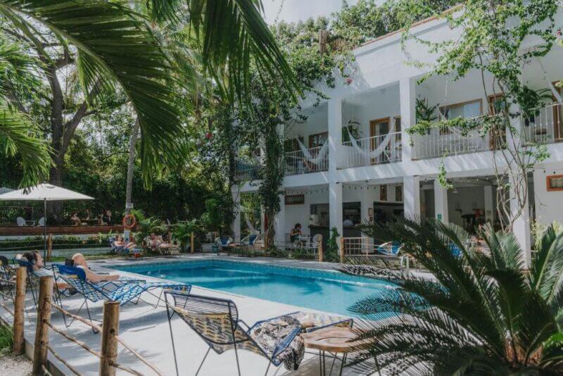 Guests unwind by the outdoor pool at a white two-story hotel with balconies, surrounded by tropical plants. A palomino sculpture and vibrant pavo real decor add unique charm as people lounge in this relaxing oasis.