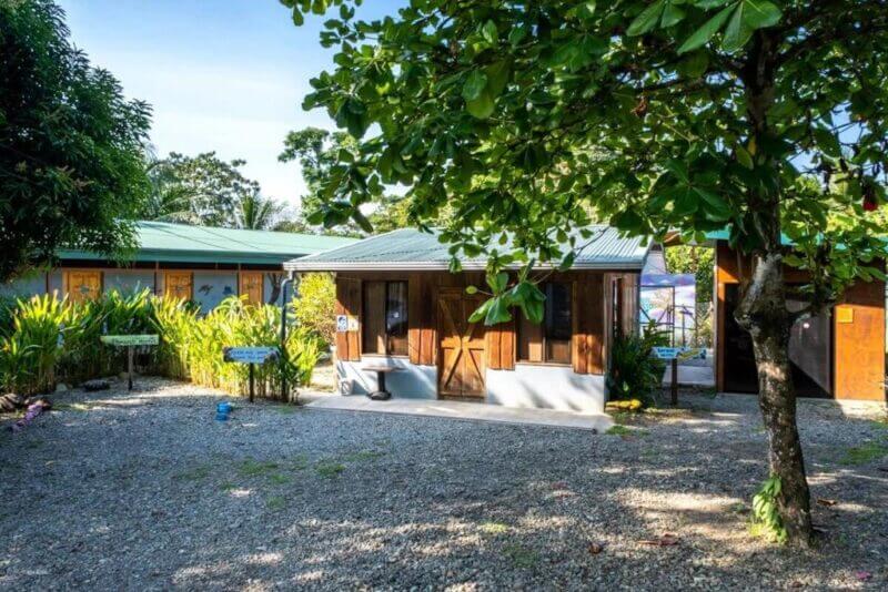 A small wooden building with a tin roof stands in a garden area with trees, greenery, and a gravel-covered ground under a clear sky—perfect for those seeking cozy Uvita accommodation.