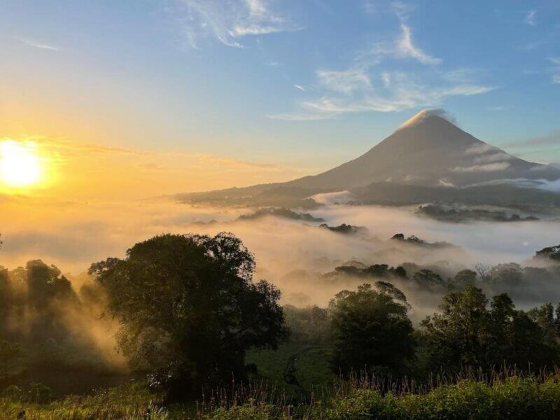 Sunrise over a misty landscape with dense trees in the foreground, a large conical volcano in the background, and the serene atmosphere reminiscent of Sangregado Lodge under a clear sky.