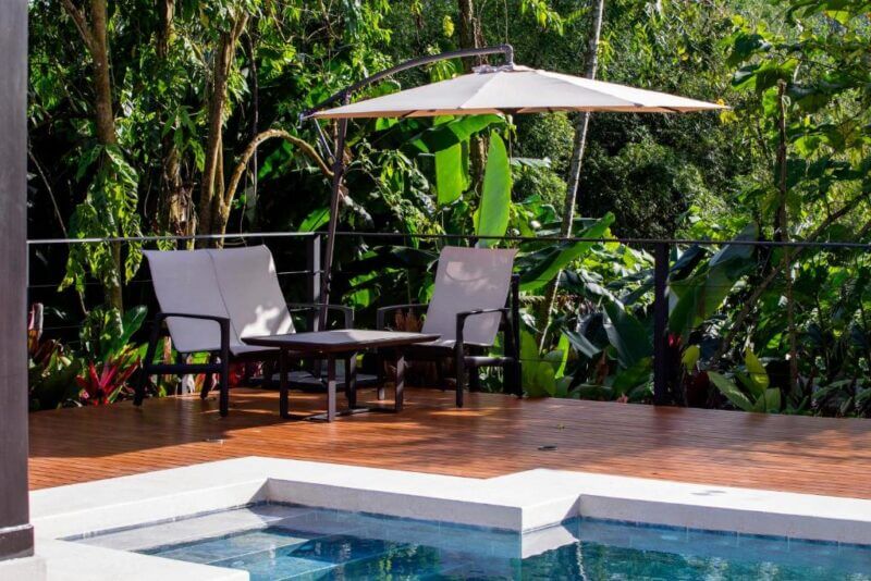 Two lounge chairs and a table sit under a large umbrella on a wooden deck beside the pool at Arenal Manoa Resort, surrounded by lush green tropical plants.