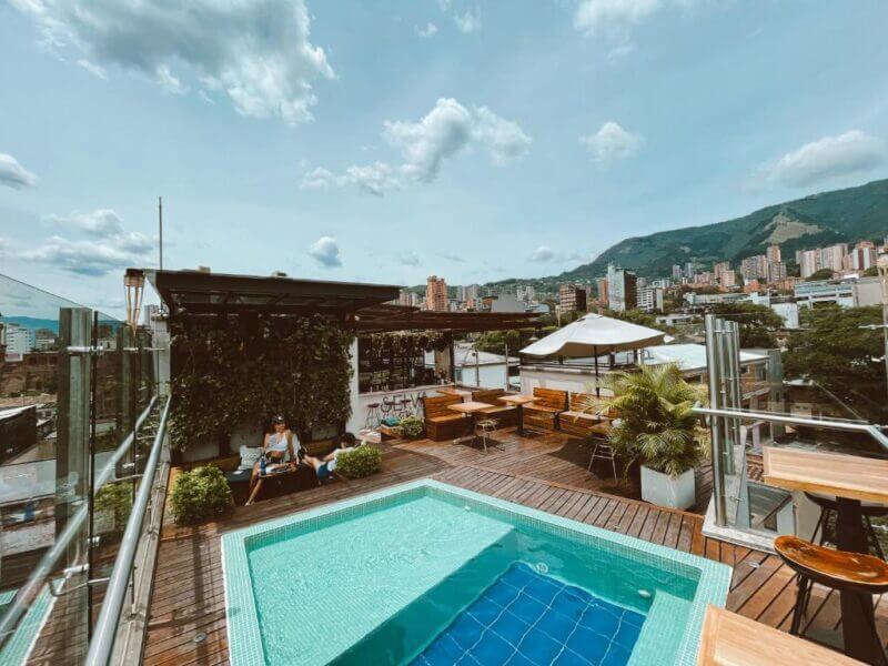 Rooftop terrace at Rango Hostel Medellin featuring a small square pool, cozy seating areas, lush plants, umbrellas, and stunning cityscape views with mountains in the background under a partly cloudy sky.