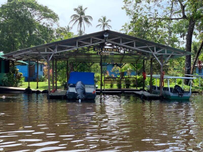 A covered dock with boats moored beneath it, displaying a sign that reads “All Rankin’s Lodge,” is nestled on a river in Tortuguero, surrounded by tropical trees and plants.