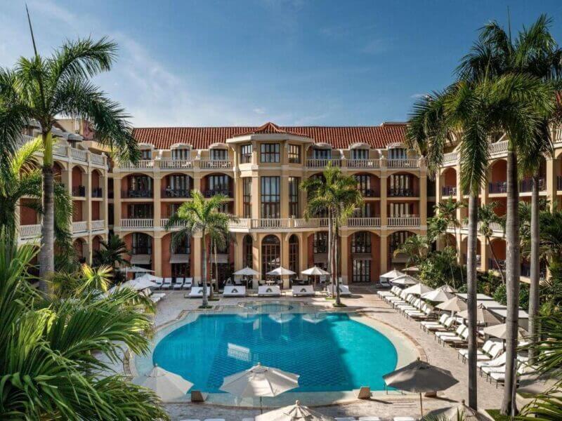 The luxury hotel Sofitel Santa Clara in Cartagena features balconies overlooking a circular outdoor swimming pool, surrounded by lounge chairs, umbrellas, and palm trees under a clear sky.